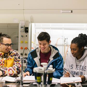 Three students stand at lab table looking at open books