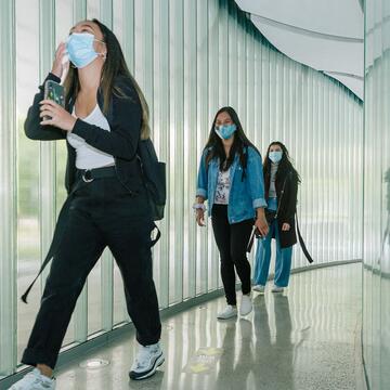 Students in masks walk along a corridor inside a building, one talking on a phone.