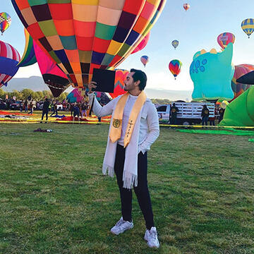 Brian Munguia holding their graduation cap and wearing their stole in front of hot air balloons.