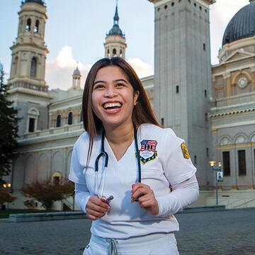 Ricci Baccay smiling in front of St. Ignatius while wearing their scrubs and holding their stethoscope.