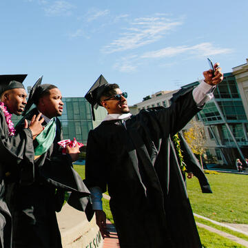 Three graduates in regalia take a selfie in front of the library.