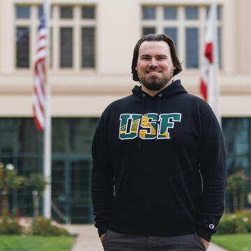 Student wearing a USF sweatshirt stands in front of a building with US and California flags flying.