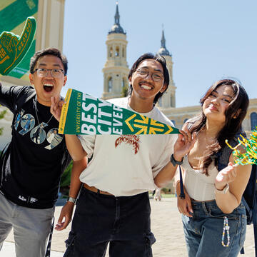 Three students cheer while holding banners, foam fingers, and pom poms