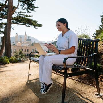 Student in nursing scrubs uses a computer on a bench.
