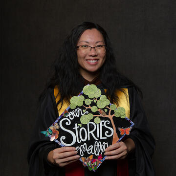 USF graduate in gown, holding cap that says "Our Stories Matter"
