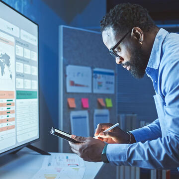 Man in business attire looking at phone and monitor screen, which displays data