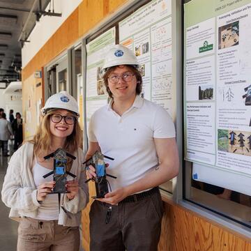 Two engineering students holding objects and standing in front of a research poster