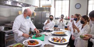 Professor in culinary uniform demonstrates on prep table in a commercial kitchen while students in culinary uniforms watch and take pictures