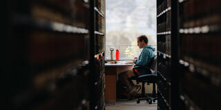 Student seen through a row of bookshelves studies at a table.