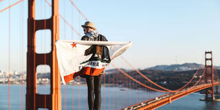 Student wrapped in a California flag stands on a high point with the Golden Gate Bridge behind.
