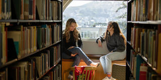 Two USF students chatting in the library, surrounding by rows of books
