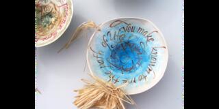 Decorative ceramic bowl with calligraphic text spiraling inward, reading “You make known to me the path of life,” and adorned with a raffia accent.