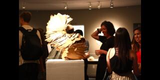 Gallery scene with people socializing around a sculptural artwork made of layered, fan-like wooden pieces on a pedestal.