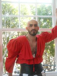 Eric Garcia in his drag persona, Churro Nomi, wearing bright red flowy top with large sleaves, large wide black belt, and wide striped black and white pants