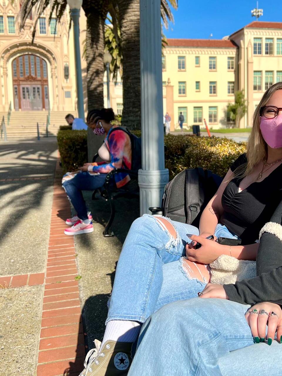 Two students sitting in the sunshine on a bench in front of Lone Mountain.