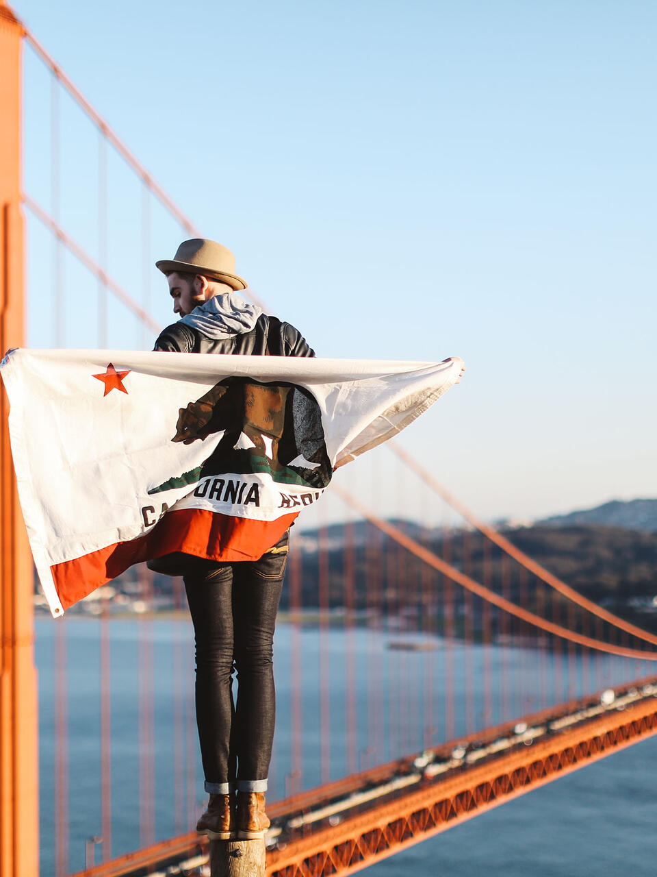 Student wrapped in a California flag stands on a high point with the Golden Gate Bridge behind.